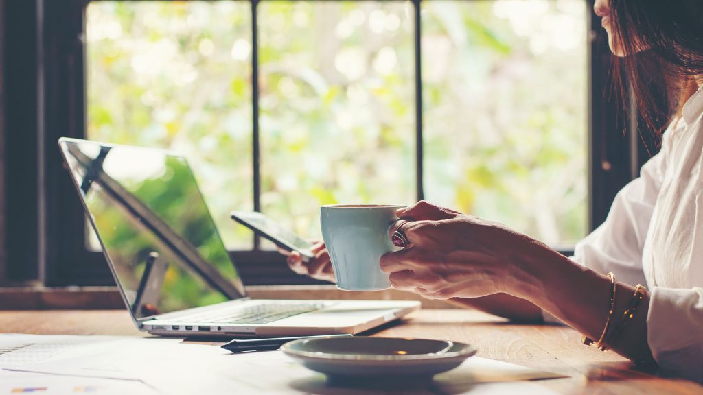 Woman working from home holding a coffe and his phone with her computer open